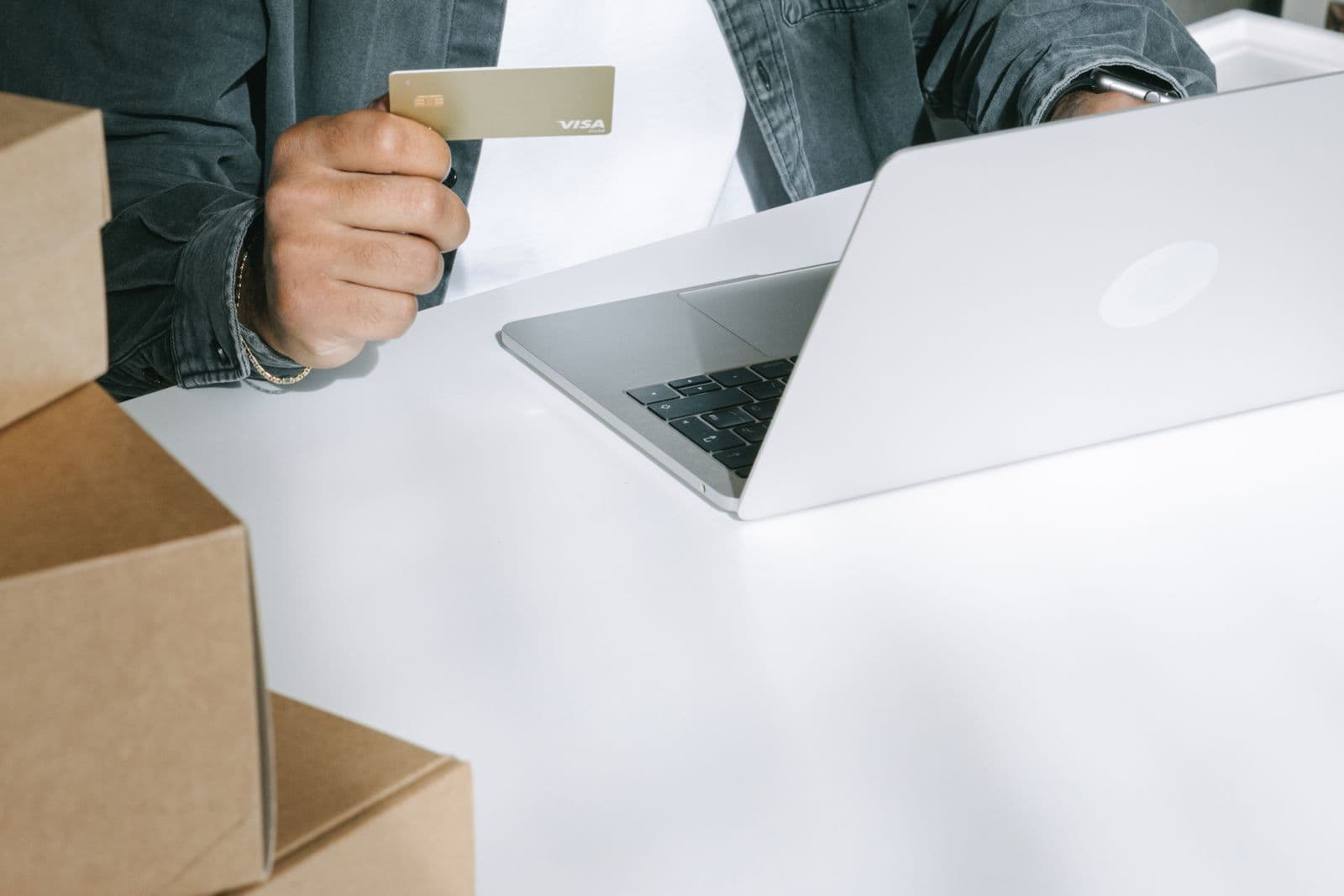 Person holds a credit card beside an open laptop, with cardboard boxes on the table.