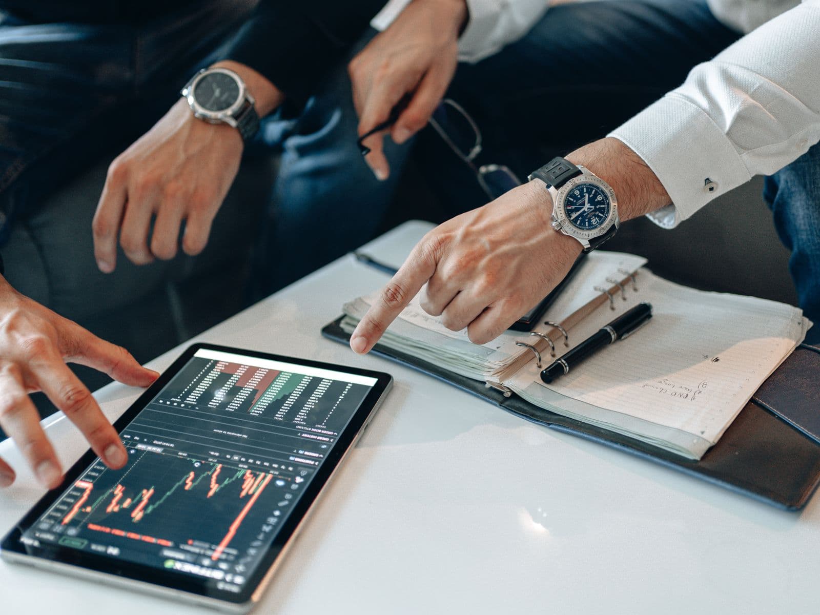 People point at a tablet with financial charts beside an open notebook and pens.