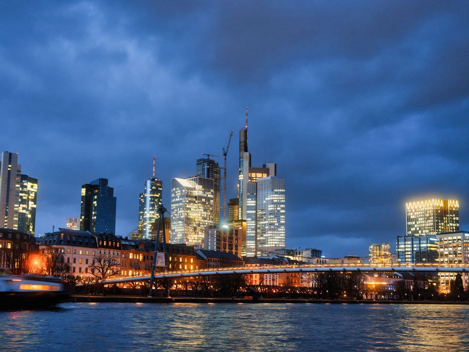Illuminated city skyline beside a river under a cloudy evening sky.